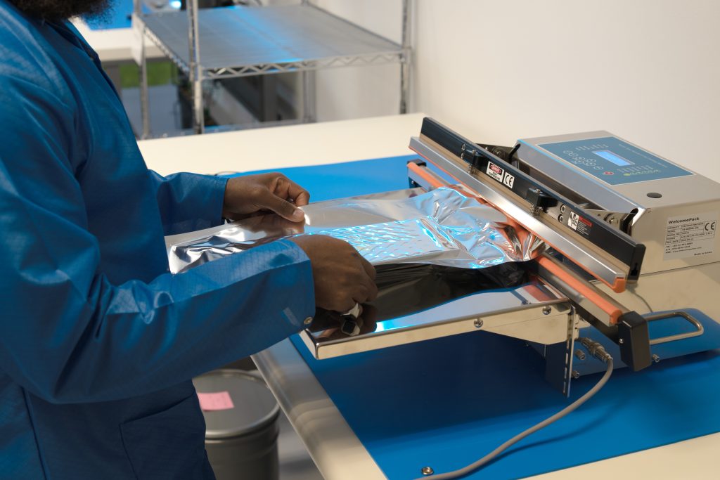 Worker in a blue uniform uses a heat-sealing machine to seal a shiny aluminum foil bag.